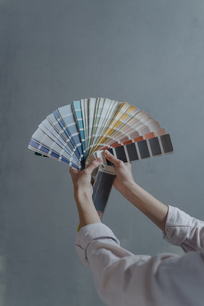 Hands displaying a fan of paint color swatches against a gray wall, ideal for home renovation projects.